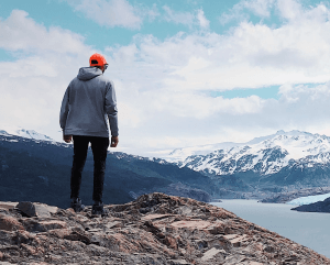 Person standing on rocky hill overlooking snowy mountains.