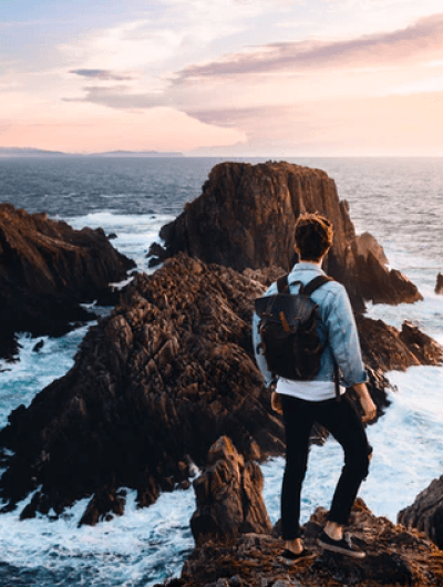 Person with backpack overlooking rocky ocean cliff.